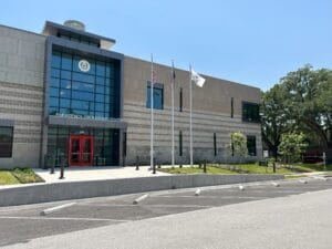 Fort Bend Emergency Operations Center A Emergency Operations Center building with large glass entrance, red double doors, multiple flagpoles, and a landscaped front area under a clear blue sky.