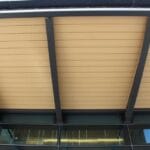 Upward view of a building overhang with tan soffit panels supported by black metal beams above a row of glass windows.